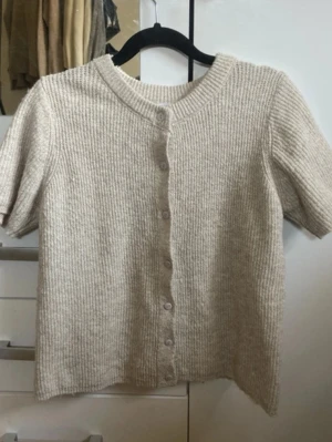 Beige ribbad kortärmad kofta - Kortärmad kofta i beige ribbstickat material med rund halsringning och knappar framtill. Mjuk och enkel design som passar perfekt till lager på lager. Ingen synlig logga eller märke.