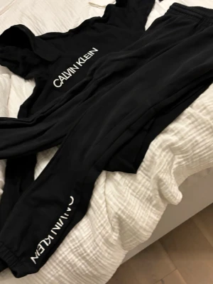 Svart loungewear-set Calvin Klein - Snyggt svart loungewear-set från Calvin Klein med tydliga logotryck på både tröja och byxor. Setet består av en mjuk långärmad tröja och matchande byxor med resår i midjan. Perfekt för chill dagar hemma. Materialet känns mjukt och bekvämt mot huden. Bra skick hör av er vid mer information storlek 14years!