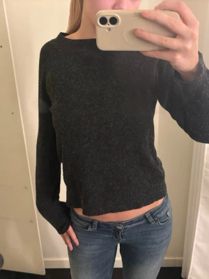Mörkgrå stickad tröja - Mysig mörkgrå stickad tröja med rund halsringning och långa ärmar. Tröjan har en något kortare, rak passform och är perfekt att matcha med jeans. Materialet ser ut att vara mjukt och bekvämt, perfekt för kyligare dagar.