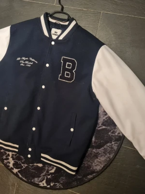 Mörkblå bomberjacka med vita ärmar - Snygg mörkblå bomberjacka från New Canner med vita ärmar och broderad text på bröstet. Stor bokstav 'B' i tyg på vänster sida. Jackan har ribbade muddar med vita ränder och tryckknappar framtill. Klassisk college-stil som passar perfekt till streetwear.