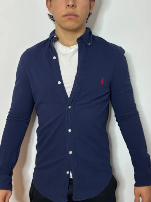 Mörkblå skjorta från Ralph Lauren - Snygg mörkblå skjorta från Ralph Lauren med klassisk krage och knappar framtill. Skjortan har lång ärm och den ikoniska röda loggan broderad på bröstet. Perfekt för en stilren och avslappnad look.