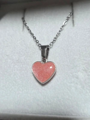Halsband med hjärthänge som lyser - Snyggt halsband med ett hjärtformat hänge i rosa som skimrar i ljuset och lyser svagt lila i mörkret. Kedjan är i silverfärgad rostfritt stål och hänget har en slät, glansig yta. Perfekt för dig som gillar unika detaljer och vill sticka ut lite extra. Hänget är handgjort och arbetat i lager samt monterat på kedja. Vanligt dagsljus räcker för att hänget ska ändra färg till lila i mörkare omgivning. Kedjan är 45 cm i rostfritt stål. (Tål vatten utan att missfärgas).