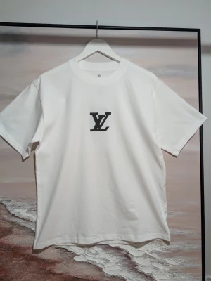 Iouis Vuitton T-shirt  - Klassisk vit t-shirt med svarta 3D-detaljer Stilren vit t-shirt i mjuk bomull med kontrasterande detaljer. Tröjan har en centrerad logotyp på bröstet och ett större grafiskt tryck på ryggen. Alla tryck är utförda i ett upphöjt, svart 3D-material som ger plagget en exklusiv textur och djup. Färg: Vit med svarta detaljer. Design: Klassisk passform, rundhalsad och korta ärmar. Detalj: Taktilt 3D-tryck för en modern look.