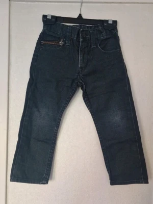 Grå/svarta jeans med dragkedjedetalj - Snygga mörkgrå jeansbyxor med klassisk femficksdesign och en cool dragkedjedetalj på ena fickan. Byxorna har resår i midjan för extra komfort och raka ben. Inga funna skavanker utöver att det finns slitage på  höger bens fåll (se bild) samt att färgen är blekt på knäna. Stl 98