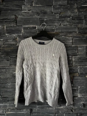 Grå kabelstickad tröja Polo Ralph Lauren - Kabelstickad tröja i ljusgrått från Polo Ralph Lauren. Klassisk rund halsringning, ribbade muddar och nederkant samt diskret broderad logga på bröstet. Perfekt för dig som gillar stilrena och tidlösa plagg i mjukt material.