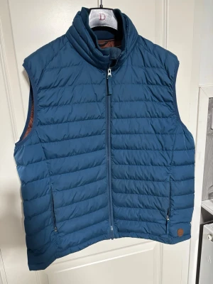 Blå dunväst från Dressmann - Snygg blå dunväst från Dressmann med hög krage och dragkedja framtill. Västen har två fickor med dragkedja och quiltad design. Perfekt för lager-på-lager och ger en sportig look. Insidan har en kontrastfärg i brunt. Passar till många olika stilar.