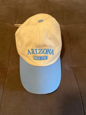 Beige och blå Arizona keps - Snygg keps i beige med ljusblå skärm och broderad text 'ARIZONA EST. 1751' framtill i blått. Klassisk form med justerbar rem bak. Perfekt för dig som vill ha en stilren och trendig accessoar till vardagslooken.