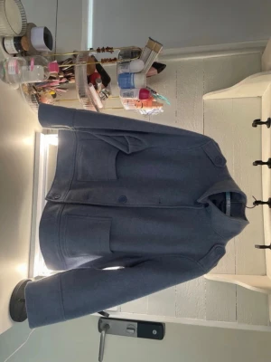 Blå jacka  - Snygg blå jacka i ullmaterial med raka linjer och stora framfickor. Jackan har hög krage, knäppning framtill med stora knappar och en minimalistisk design. Perfekt för dig som gillar stilrena och klassiska plagg.