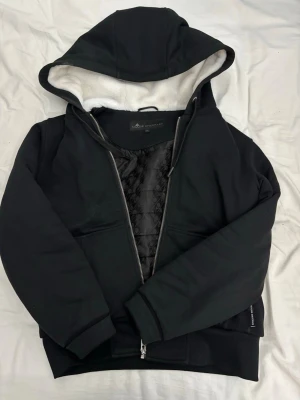 Svart hoodie från Moose Knuckles - Säljer en svart hoodie från Moose Knuckles med dragkedja, stor huva och vit fleecefoder på insidan av huvan. Hoodien har ett snyggt quiltat innerfoder och en ikonisk logga på ärmen. Perfekt för dig som vill ha en stilren och varm hoodie. Passar dig runt 168-177. Pris kan diskuteras på snabb affär