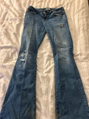Blå bootcut jeans från LTB - Säljer ett par blå bootcut jeans från LTB, modell Roxy. Jeansen har slitna detaljer och hål på låret och knät för en trendig look. Klassisk femficksmodell med dragkedja och knapp. Perfekta för dig som gillar en avslappnad och cool stil. Se sista bilderna för hur det ser ut längst ner på byxorna då dom är slitna där 
