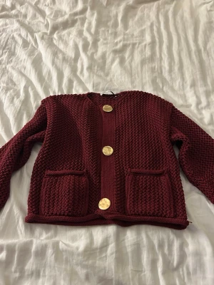 Mörkröd stickad kofta med guldknappar - Snygg mörkröd stickad kofta med grov struktur och tre stora guldknappar framtill. Koftan har två framfickor och rund halsringning.