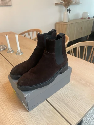 Bruna Chelsea boots i mocka från Vagabond - Snygga bruna Chelsea boots i mocka med klassiska elastiska paneler på sidorna och svart sula. Perfekt för dig som vill ha en stilren och tidlös look. Bootsens form är smal och elegant, och de är lätta att dra på tack vare resårdetaljerna.