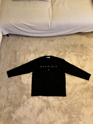 Svart långärmad topp Margiela  - Svart långärmad topp från Margiela 6 med rund halsringning. Framsidan har texten 'MARGIELA 6' i små pärlor som ger en glittrig effekt. Tillverkad i mjukt material, perfekt för en clean och stilren look.