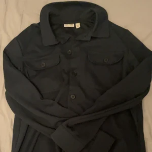 Mörkblå overshirt från Only&sons - Stilren mörkblå overshirt från only&sons med klassisk krage och stora bröstfickor med lock och knapp. Overshirten har knäppning framtill och är gjord i ett mjukt tyg som känns bekvämt. Perfekt att bära öppen eller stängd för en avslappnad look. Använd 1 gång