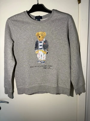 Grå sweatshirt Polo Bear Ralph Lauren - Grå sweatshirt från Ralph Lauren med det ikoniska Polo Bear-motivet på bröstet. Tröjan har rund halsringning och ribbade muddar vid ärmslut och nederkant. Perfekt för en avslappnad och stilren look. Nypris: 1999kr!