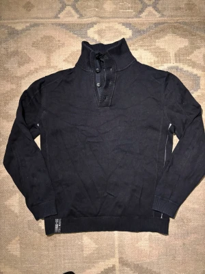 Svart stickad tröja med hög krage - Svart stickad tröja med hög ribbad krage och knappslå framtill. Tröjan har långa ärmar och ribbade muddar vid ärmslut och nederkant. Perfekt för dig som gillar en stilren och enkel look.