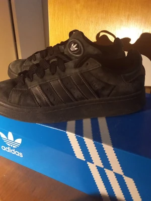 Svarta Adidas Campus  sneakers - Snygga helsvarta Adidas  campus sneakers i mocka med klassiska tre ränder på sidan och rund tå. Skorna har svart sula, svarta skosnören och Adidas-logga på plösen. Perfekta för dig som gillar stilrena och tidlösa sneakers. Pris kan diskuteras.