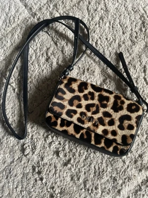 Leopardmönstrad axelväska från DKNY - Riktig axelväska från DKNY med leopardmönstrad framsida och svarta detaljer. Väskan har justerbar axelrem och ett huvudfack med innerficka. Perfekt för att lyfta vilken outfit som helst med lite extra attityd.