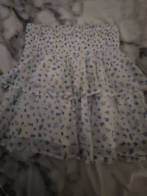 Vit blommig volangkjol - Supersöt kort kjol i vitt med blått blommönster och volanger. Kjolen har bred smock i midjan för skön passform och är gjord i ett lätt, luftigt material som passar perfekt till varma dagar. Perfekt för dig som gillar romantisk och trendig stil.