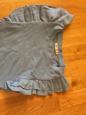 Blå ribbad topp med volang från Zara - Supersöt ljusblå topp från Zara med ribbad struktur och volangdetaljer på ärmarna. Perfekt att styla med jeans eller kjol för en trendig look. Mjuk och skön, passar dig som gillar feminina detaljer.