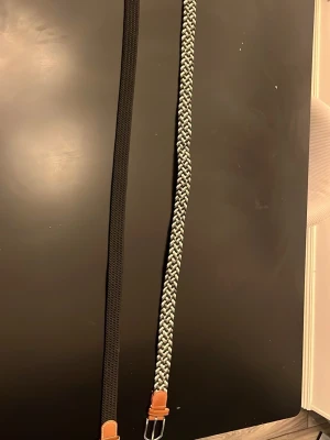 Två flätade bälten i svart och grått - Säljer två stilrena flätade bälten, ett helsvart och ett i svart och grått mönster. Båda har detaljer i brunt skinn vid spännet och passar perfekt till jeans eller chinos. Snygga accessoarer för att lyfta din outfit.