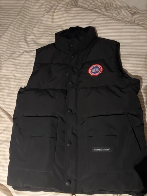 Svart väst Canada Goose - Snygg svart dunväst från Canada Goose med hög krage och tryckknappar framtill. Västen har två stora fickor och klassisk Canada Goose-logga på bröstet. Perfekt för lager-på-lager och håller dig varm under kyliga dagar.