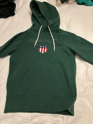 Grön hoodie från GANT - Mörkgrön hoodie från GANT i mjukt bomullsmaterial. Klassisk modell med huva och ribbade muddar vid ärmslut och nederkant. Perfekt för en chill och avslappnad stil. Ny skick och storlek S. Skriv vid frågor eller fler bilder. 