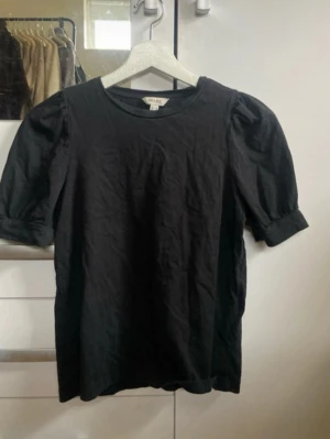 Svart puffärmad topp från Weekday - En svart topp från Weekday med puffärmar och rund halsringning. Toppen har en enkel, stilren design och är gjord i mjuk bomull. Perfekt för dig som gillar minimalistisk stil med en twist.