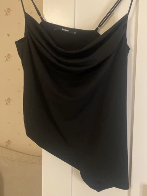 Svart linnetopp från Bik Bok - Svart linnetopp från Bik Bok med tunna axelband och snygg draperad framsida. Toppen har asymmetrisk nederkant och är tillverkad i ett mjukt syntetmaterial som känns skönt mot huden. Perfekt för dig som gillar stilrena och trendiga plagg.