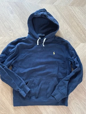 Mörkblå hoodie från Polo Ralph Lauren - Snygg mörkblå hoodie från Polo Ralph Lauren med klassisk gul broderad logga på bröstet. Hoodien har vit dragsko i huvan och en stor magficka framtill. Tillverkad i mjukt bomullsmaterial och har ribbade muddar vid ärmslut och nederkant.