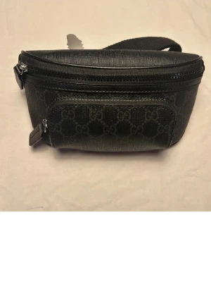 Svart GG magväska - Snygg svart magväska med klassiskt GG-mönster i mörkgrått. Väskan har två dragkedjefack och justerbar rem. Tillverkad i tåligt läderliknande material med detaljer i svart. Perfekt för dig som vill ha en stilren och trendig accessoar. Har slängt det gröna paketet då det tog mycket onödig plats.