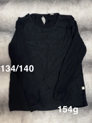 Svart långärmad topp med struktur - Svart långärmad topp med diskret prickig struktur på framsidan och axelparti. Rund halsringning med knappdetalj bak i nacken. perfekt för lager på lager eller som den är.🌸 Fint använt skick. Storlek 134/140