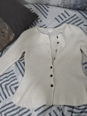 Vit ribbstickad kofta från H&M - Säljer en vit ribbstickad kofta från H&M med rund halsringning och bruna knappar framtill. Koftan har lång ärm och är lätt figursydd för en snygg passform. Perfekt att slänga över en topp när det är kyligt ute plus den är helt ny som ni ser på storleken så står det L men när man testar den så är det typ s/xs ☺️