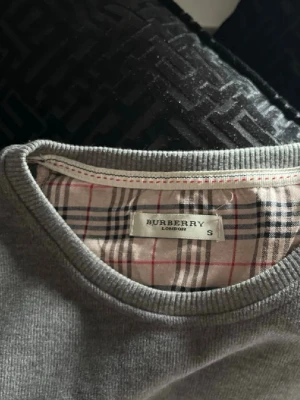 Grå långärmad topp från Burberry - Snygg grå långärmad topp från Burberry med klassiskt rutigt foder i halsen och diskret Burberry-logga broderad på bröstet. Rund halsringning och mjukt material som känns skönt mot huden. Perfekt för dig som gillar stilrena plagg med exklusiv känsla.