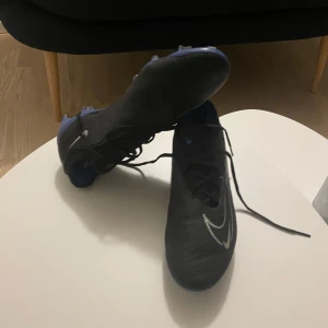 Nike Phantom GT fotbollsskor svart/blå - Säljer ett par Nike Phantom GT fotbollsskor i svart med blå detaljer och vit Nike-logga. Skorna har snörning och är designade för bra grepp på planen. Ovandelen är i syntetmaterial för optimal bollkontroll och komfort. Perfekta för dig som vill ta ditt spel till nästa nivå. Nästan aldrig använda köpta efter en skada och sedan slutade jag spela.