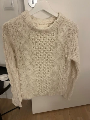 Vit kabelstickad tröja - Säljer en krämvit kabelstickad tröja med grov struktur och dekorativa bollar längs flätmönstret. Tröjan har rund halsringning och långa ärmar. Perfekt för kalla dagar och riktigt mysig att bära.