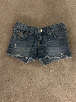 Blå jeansshorts med rå kant - Snygga blå jeansshorts med rå, fransig kant och slitna detaljer. Klassisk femficksmodell med knapp och dragkedja framtill samt dekorativa bakfickor med knapp. Perfekta för varma dagar och en avslappnad stil.