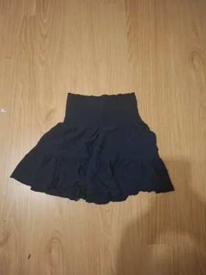 Svart volangkjol med resårmidja - Kort svart kjol med bred resår i midjan och volangdetaljer. Kjolen har en luftig och flowy passform som ger en avslappnad look. Perfekt att matcha med en enkel topp eller hoodie.