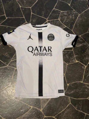 PSG vit matchtröja Jordan x Nike - Säljer en vit PSG matchtröja med svart vertikal rand, Jordan-logga och Paris Saint-Germain-märke på bröstet. Tröjan har svarta detaljer vid ärmslut och sponsortryck från Qatar Airways och Ligue 1 Uber Eats. Tillverkad i lätt funktionsmaterial, perfekt för fotboll.