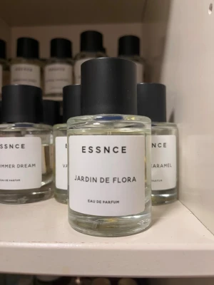 ESSNCE Jardin de Flora parfym - Minimalistisk parfymflaska från ESSNCE med doften Jardin de Flora. Flaskan är cylindrisk i genomskinligt glas med svart matt kork och vit etikett med svart text. Innehållet är ljust gult. Perfekt för dig som gillar fräscha och blommiga dofter.