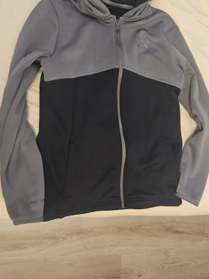 Grå och svart hoodie från Under Armour - Snygg hoodie från Under Armour i grått och svart med dragkedja framtill. Tillverkad i ColdGear-material som håller dig varm och bekväm. Hoodien har huva och två sidofickor, perfekt för chill dagar eller träning.