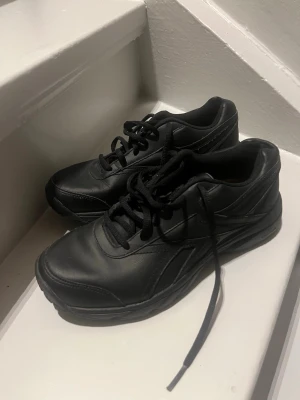 Svarta Reebok sneakers i läder - Snygga helsvarta sneakers från Reebok i läder med klassisk design och slitstark yttersula som är både olje- och halkresistent. Skorna har lågt skaft, svarta skosnören och diskreta detaljer längs sidorna. Perfekta för dig som gillar stilrena och funktionella sneakers.