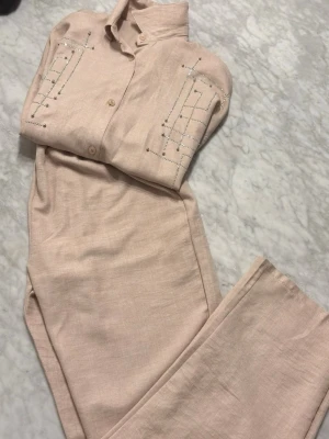 Beige set  med dekorativa detaljer - Säljer en beige set med krage och knappar framtill. Det har dekorativa silverfärgade nitar och linjer på bröstet som ger ett coolt och unikt uttryck. Materialet är mjukt och lätt, perfekt för dig som vill ha något stilrent men ändå sticker ut.