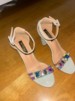 Glittriga pumps med färgglada stenar - Säljer ett par glittriga pumps från SOPHIA'S med bred klack och öppen tå. Skorna har en rem runt ankeln och en transparent rem över tårna dekorerad med stora färgglada stenar i olika former. Perfekta för dig som vill sticka ut och älskar bling!