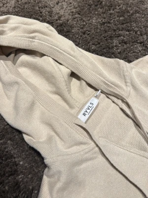 Beige hoodie med dragkedja från RAVLS - Snygg beige hoodie från RAVLS med dragkedja framtill och två fickor. Tillverkad i ett mjukt, tunt material som känns skönt mot huden. Klassisk huva med dragsko och ribbade muddar vid ärmslut och nederkant. Perfekt för en avslappnad stil.