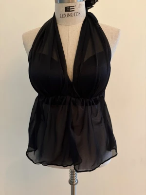 Svart topp halterneck i mesh - Svart halterneck topp i mesh med knytband i nacken och omlott framtill. Inbyggd BH med öppning i nacken. Toppen är ärmlös och har ett lätt transparent, flödande tyg som ger en luftig och trendig look. 