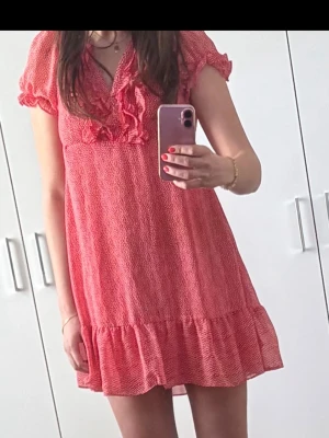 Röd kort volangklänning med v-ringning - Söt röd kortklänning med volangdetaljer och v-ringning. Klänningen har korta puffärmar och en lös passform som ger en avslappnad look. Tillverkad i ett lätt material, perfekt för varma dagar.