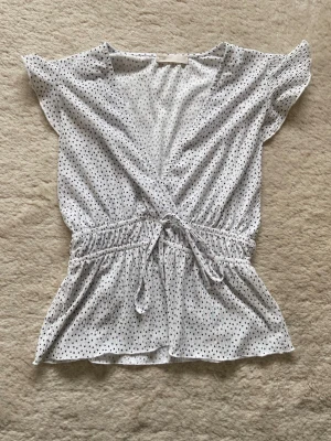Vit prickig topp - Söt vit topp med svarta prickar. Toppen har korta ärmar och volangkant nertill. Perfekt för varma dagar och enkel att styla med jeans eller kjol.