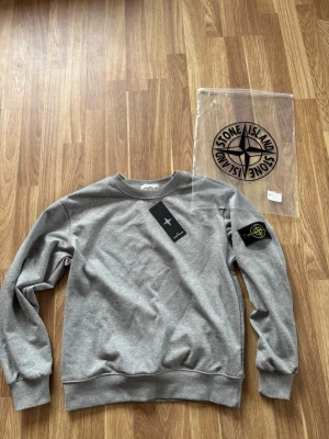 Grå stone Island tröja - Snygg grå sweatshirt från Stone Island med klassisk rund hals och ribbade muddar. Tröjan har den ikoniska svarta patchen med gul och grön logga på vänster ärm. Tillverkad i mjuk bomull för en bekväm känsla. Perfekt för en avslappnad streetwear-look.