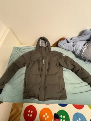 Mörkgrön pufferjacka med huva - Snygg mörkgrön pufferjacka med huva och flera dragkedjeförsedda fickor framtill. Jackan har en patch på ena ärmen och quiltad design för extra värme. Perfekt för kalla vinterdagar och har en modern, sportig look.